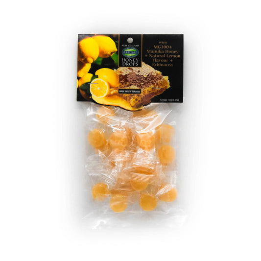 packet of Manuka Honey drops on a white background. The packaging is clear so you can see the drops but there is also a cardboard header that is black with photos of lemons including one sliced open on top of Manuka Honey comb and some text 